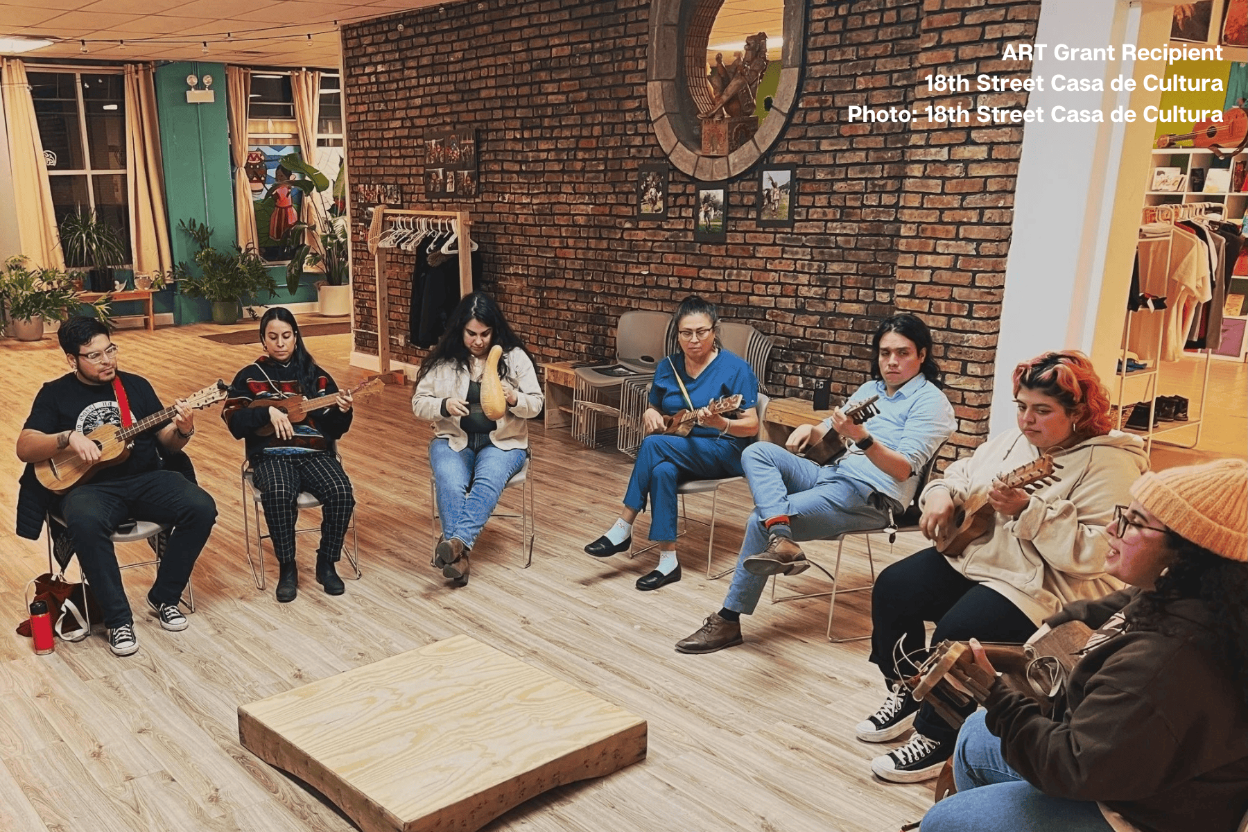 Seven students seated in circle on wooden floor learning ukuleles and traditional string instruments at ART grant recipient 18th Street Casa de Cultura in Pilsen, in welcoming cultural space with exposed brick walls, wooden sculpture, plants, and natural light from large windows